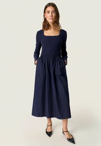 Marinblå klänning med ribbat liv, fyrkantig halsringning och en draperad kjol med fickor. Matchad med svarta spetsiga ballerinas.