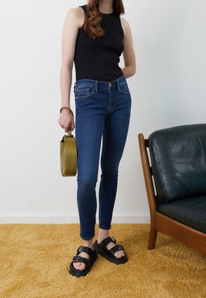 Jean skinny en denim bleu avec une taille haute, associé à un débardeur noir et des sandales compensées. Tenant un petit sac à main vert olive.