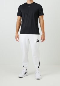 Černé sportovní tričko s krátkým rukávem, kombinované s bílými tepláky se černými postranními pruhy a logem Adidas. Viditelné bílé tenisky.