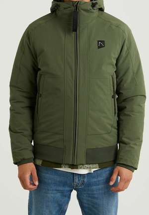 Homme portant une veste matelassée verte avec fermeture éclair frontale, poignets côtelés et poches, associée à un jean bleu sur fond uni.
