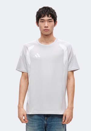 Fiatal férfi, rövid sötét hajjal, világosszürke Adidas pólót és kék farmert visel, egyszínű háttér előtt.