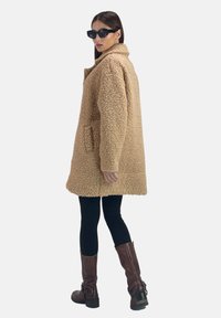 Cappotto in pelliccia sintetica beige con collo, tasche laterali e taglio dritto. Abbinato a leggings neri e stivali marroni al ginocchio.