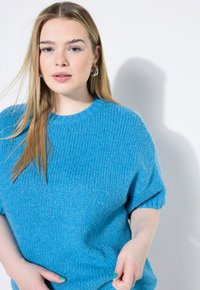 Blå stickad tröja med korta ärmar, rundad hals och ribbad textur. Händerna vilande på höfterna, mot en enfärgad vit bakgrund.