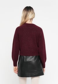 Femme aux longs cheveux blonds portant un pull bordeaux et une jupe en cuir noire, vue de dos sur un fond blanc uni.
