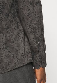 Chemise à motif sombre présentant un tissu texturé avec un design tacheté subtil, et un détail de manche boutonnée.