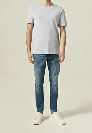 Mann trägt ein schlichtes hellgraues kurzärmeliges T-Shirt, schlanke blaue Jeans und weiße Turnschuhe, steht vor einem neutralen Hintergrund.