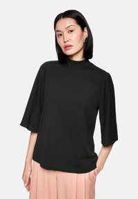 Black, mock-neck top with flared short sleeves; smooth fabric texture; features a relaxed fit, worn with light pink pleated pants.