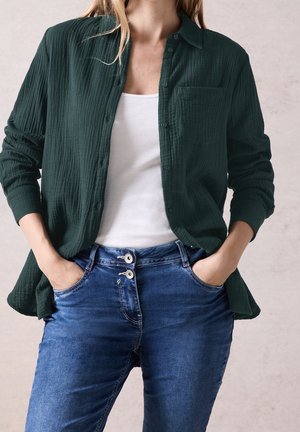 Femme portant une chemise boutonnée vert foncé par-dessus un débardeur blanc et un jean bleu à double bouton, les mains dans les poches, debout devant un fond neutre.