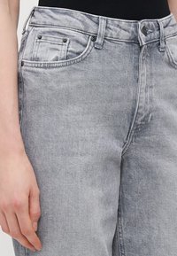 Sivi traperice s klasičnim dizajnom od pet džepova, blago istrošenim završetkom i metalnim detaljima na struku i džepovima.