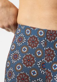 Main ajustant la taille d'un short bleu avec un motif floral et géométrique rouge, blanc et marron sur une peau nue.