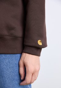 Brown sweatshirt with ribbed cuffs, featuring a small yellow logo on the left sleeve. Worn over blue denim, showcasing fabric texture.