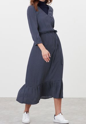 Femme portant une robe midi manches longues bleu marine à pois blancs, associée à des baskets blanches, debout sur un sol gris.