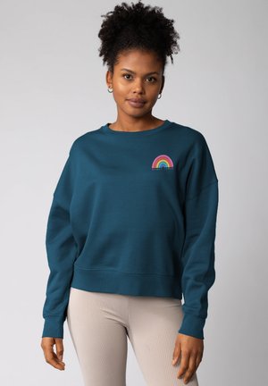 Tegen sweatshirt in teal met een relaxte pasvorm, ribbelboord rond de hals en geborduurd regenboogontwerp. Hoogwaardige stof met een zachte textuur.
