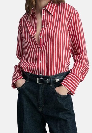 Person iført en rød og hvid lodret striped skjorte stukket ind i mørke jeans med sort bælte, hænderne puttet i forlommerne.