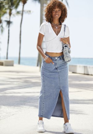 Weißes geripptes Crop-Top mit einem tiefen V-Ausschnitt, kombiniert mit einem hochgeschnittenen Jeans-Maxikleid mit einem Schlitz vorne, getragen mit weißen Turnschuhen.