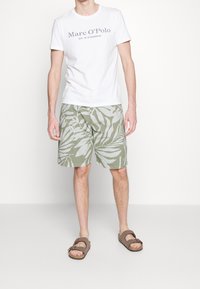 Hvid t-shirt med "Marc O'Polo" logo, grønne shorts med hvide bladmønstre og brune sandaler med stropdesign.