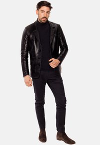 Blazer de cuero negro con tres botones, solapas acampanadas, llevado sobre un cuello alto de punto oscuro; combinado con pantalones oscuros de textura y botas de ante marrones.