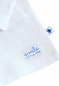 Bílá bavlněná košile s modrým vyšitým štítkem, na kterém je napsáno "smile LITTLE ONE", a malou modrou hvězdičkovou nálepkou na boku.
