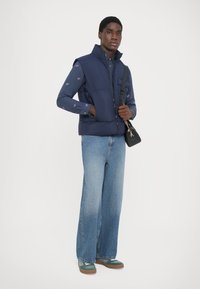 Navy puffer vest with high collar, worn over a patterned button-up shirt. Wide-leg light blue denim jeans and green-accented shoes complete the look.