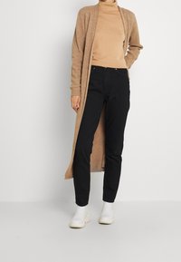 Lungo cardigan beige su un dolcevita marrone chiaro, abbinato a jeans neri slim-fit e stivaletti bianchi, su uno sfondo semplice.