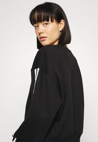 Melna pārāk liela izmēra džemperis ar garām piedurknēm, kas rotāts ar baltām svītrām uz pleciem un apaļu kakla izgriezumu. Mīksta auduma tekstūra.