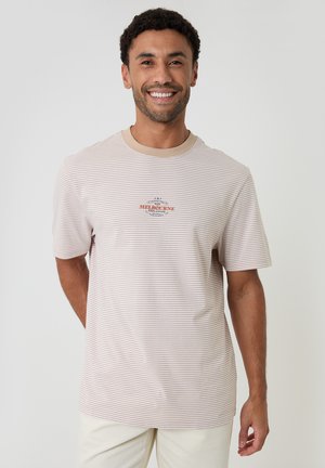 Uomo sorridente che indossa una maglietta beige e bianca a righe con la scritta "Melbourne Esplanade", in piedi con la mano dietro la schiena su sfondo semplice.