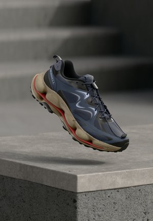 Zapatilla deportiva azul y negra con suela beige y detalles rojos, suspendida en el aire sobre un borde de cemento con escaleras al fondo.