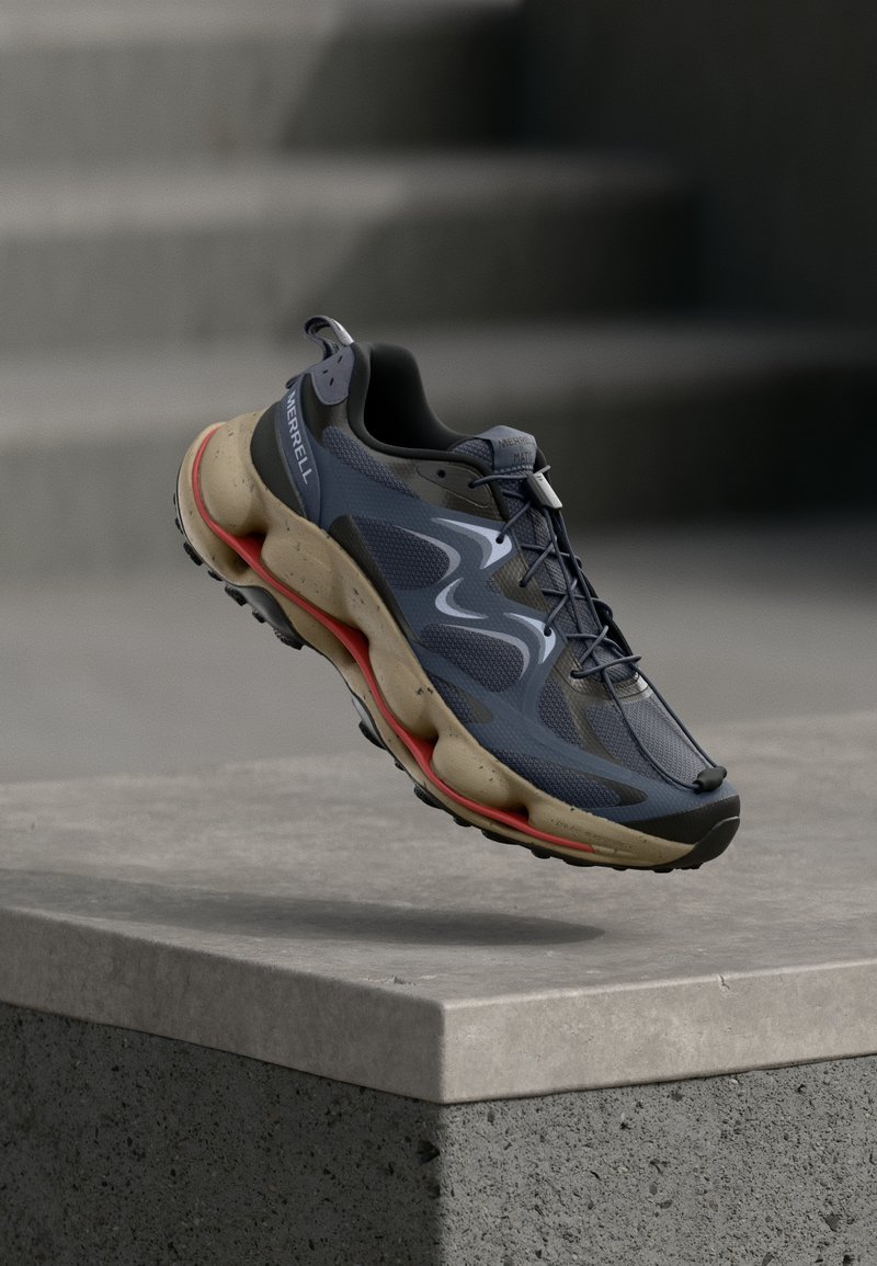 Niebiesko-czarny sportowy but z beżową podeszwą i czerwonymi akcentami, zawieszony w powietrzu nad betonowym krawężnikiem, w tle schody.