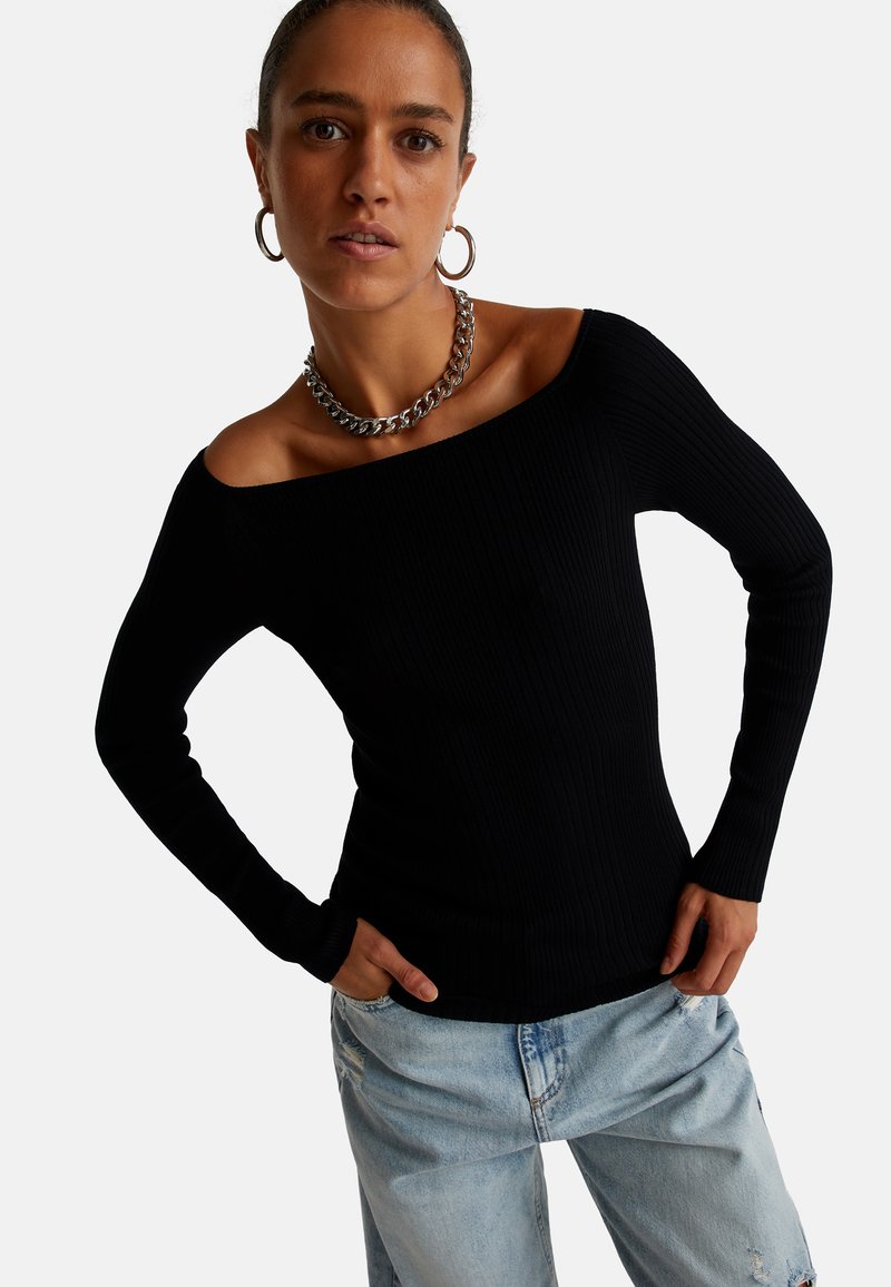 Top negro de canalé sin hombros con mangas largas, que presenta un diseño ajustado. La modelo lleva un collar de cadena plateado y pantalones de mezclilla claros.
