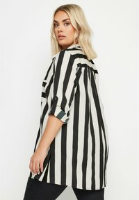 Camicia a strisce bianco e nere con colletto, maniche arrotolate e fessure laterali. Realizzata in tessuto leggero con una consistenza liscia.