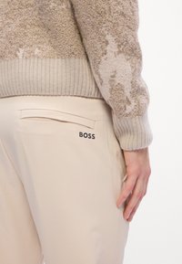 Beige broek met achterzak en zwart "BOSS" logo op de heup, gecombineerd met een gestructureerde beige gebreide trui en een ontspannen hand aan de zijkant.