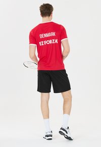 Mies, joka on pukeutunut punaiseen 'Denmark FZ Forza' -paitaan, mustiin shortseihin ja valkoisiin tennareihin, pitäen kädessään sulkapallomailaa, seisoo valkoista taustaa vasten.