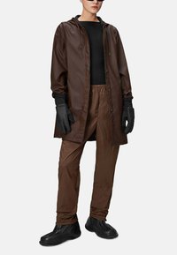 Cappotto lungo impermeabile marrone con cappuccio, abbinato a pantaloni leggeri coordinati e guanti neri. Scarpe in gomma nere completano l'outfit.