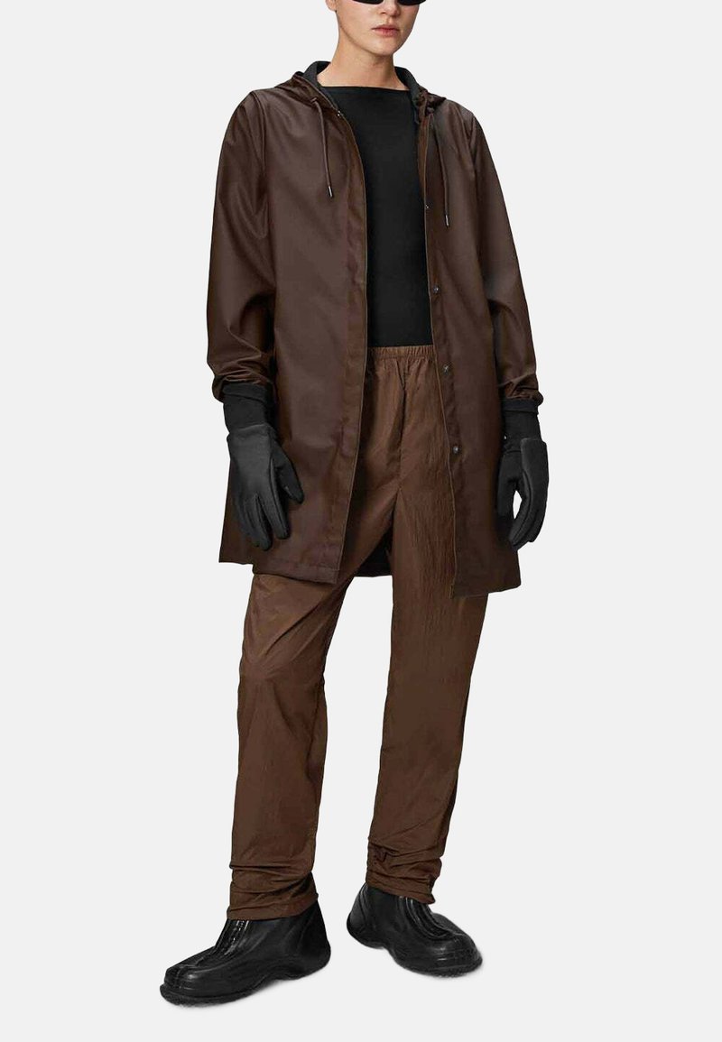 Cappotto lungo impermeabile marrone con cappuccio, abbinato a pantaloni leggeri coordinati e guanti neri. Scarpe in gomma nere completano l'outfit.