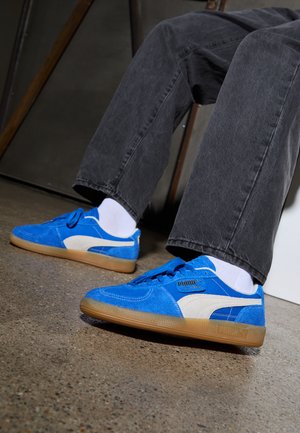 Baskets en daim bleu avec des accents blancs et une marque noire, dotées d'une semelle en caoutchouc beige. Portées avec un pantalon gris foncé et des chaussettes blanches.