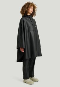 Svart vattentät poncho med hög krage, tryckknappar, framficka och vida ärmar, kombinerad med högmidjade svarta byxor och beige skor.