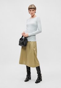 Blusa de manga longa azul clara, saia verde oliva com fenda lateral e botas de couro pretas até os joelhos, segurando uma pequena bolsa preta trançada.