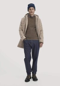 Beiger Wollmantel über einem braunen Pullover, blauen Jeans und schwarzen Schuhen. Eine marineblaue Mütze und ein kariertes Hemd verleihen dem Outfit Details.