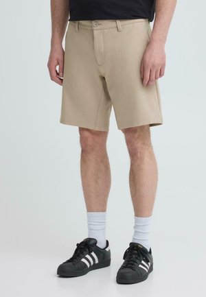 Uomo che indossa pantaloncini beige fino al ginocchio, calzini bianchi alti e sneakers Adidas nere con strisce bianche, in piedi su uno sfondo chiaro semplice.