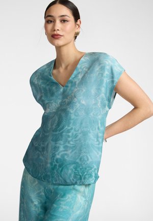 Kvinde iført en blå mønstret V-hals bluse og matchende bukser, poserer med den ene hånd på hoften foran en ensfarvet baggrund.