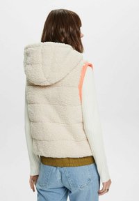 Knuffelige crèmekleurige hoodie-vest met oranje accenten, gewatteerd ontwerp en een geribbelde zoom, gecombineerd met een wit shirt met lange mouwen en lichtblauwe jeans.