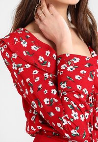La blusa floral roja presenta patrones de flores en blanco y verde, un escote cuadrado, mangas largas abullonadas y una cintura fruncida.