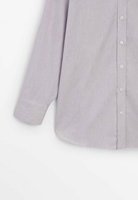 Chemise longue à manches longues de couleur violet clair avec de petits boutons blancs et des poignets larges sur un fond blanc.
