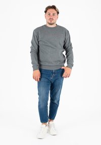 Uomo in piedi contro uno sfondo bianco, indossa un felpa grigia, jeans blu e sneakers bianche, rivolto in avanti con un'espressione neutra.