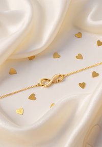 Bracciale in oro con simbolo dell'infinito e accenti scintillanti, su uno sfondo in tessuto seta crema con forme a cuore dorate.