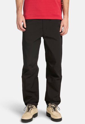 Mandlig model iført sorte bukser med lige ben, beige snørebåndssko og en rød skjorte, stående med afslappede hænder langs siden.