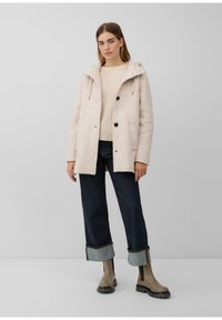 Manteau capuchon beige clair avec des boutons noirs, en tissu texturé, deux poches avant, porté sur un pull crème et un jean denim foncé, associé à des bottes claires.