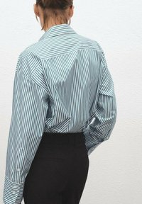 Striped shirt in green and white fabric, featuring a classic collar, long sleeves, and buttoned cuffs. Worn with black trousers.