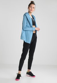 Giacca con cappuccio azzurro chiaro sopra un top corto nero, abbinata a pantaloni neri a gamba tapered e sneakers nere con dettagli rosa. Texture morbide in tutto.