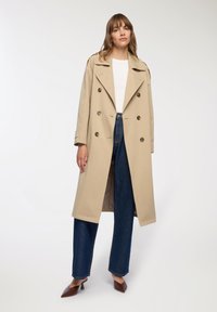 Beżowy płaszcz trencz z podwójnym zapięciem, dużymi guzikami i szerokimi klapami, w połączeniu z niebieskimi dżinsami z wysokim stanem i brązowymi butami.